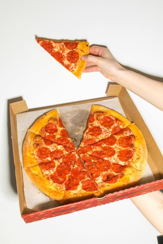 A hand holding a slice of pepperoni pizza above an open pizza box containing several slices.